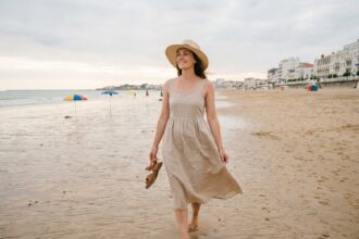 Femme se promenant pieds nus sur la plage des Sables-d'Olonne avec chapeau de paille et robe en lin, vue de dos avec la promenade en arrière-plan