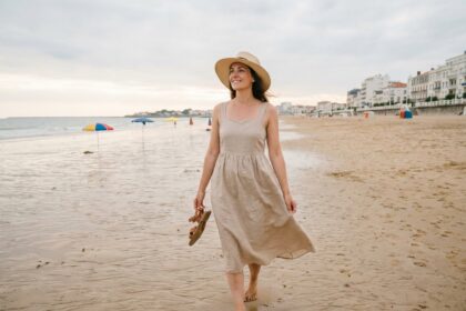 Femme se promenant pieds nus sur la plage des Sables-d'Olonne avec chapeau de paille et robe en lin, vue de dos avec la promenade en arrière-plan